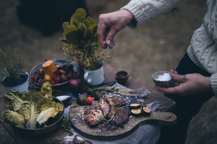 Person seasoning a spread of grilled meat and vegetables outdoors.